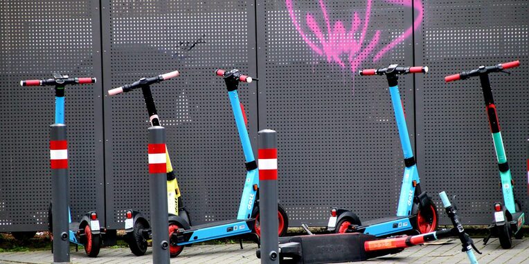 mehrere blaue E-Scooter stehen und liegen auf einem Platz vor einem grauen Metallzaun