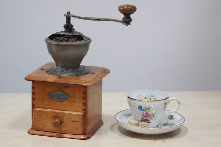 Eine alte braune Kaffeemühle zum drehen mit einer alten weiß geblümten Kaffeetasse und zwei Stückchen Würfelzucker auf dem Unterteller