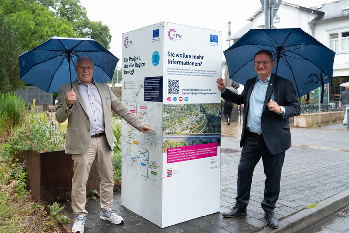 Infostele mit Horst Amann links und Bürgermeister Dirk Gene Hagelstein rechts. Beide halten einen Regenschirm.